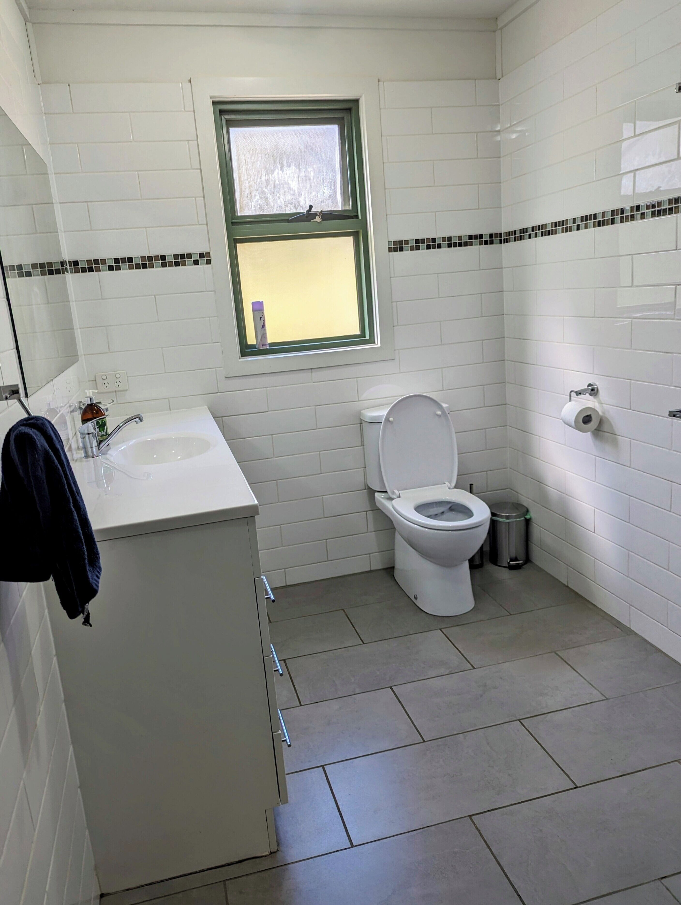 A small, clean bathroom with white tiles, a white sink, a toilet, and a window above the toilet.