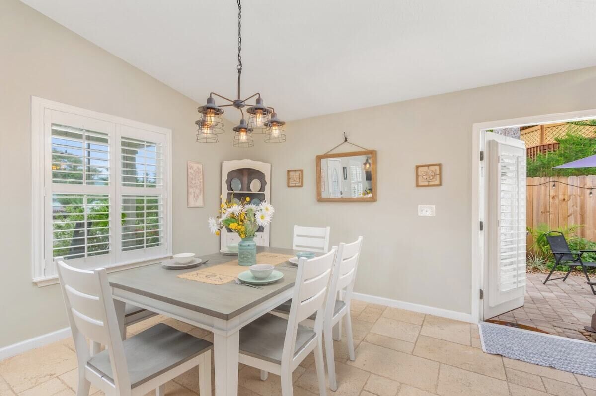 A dining area with a table set for four, white chairs, a chandelier, and a view of the backyard through a sliding glass door.