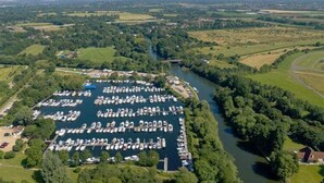 Eine Luftaufnahme eines Yachthafens mit zahlreichen Booten, die an einem Fluss liegen, umgeben von üppiger Vegetation und Feldern.