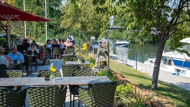 Ein Essbereich im Freien mit Tischen und Stühlen, Blick auf einen Fluss und Boote im Hintergrund.