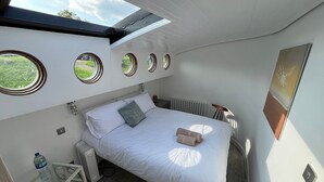 Ein gemütliches Schlafzimmer mit Dachfenster, einem Bett mit weißer Bettwäsche, einem Heizkörper und einem kleinen Tisch mit einer Wasserflasche.