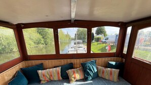 Eine hölzerne Bank mit Kissen, große Fenster und Blick auf einen Kanal mit Booten.