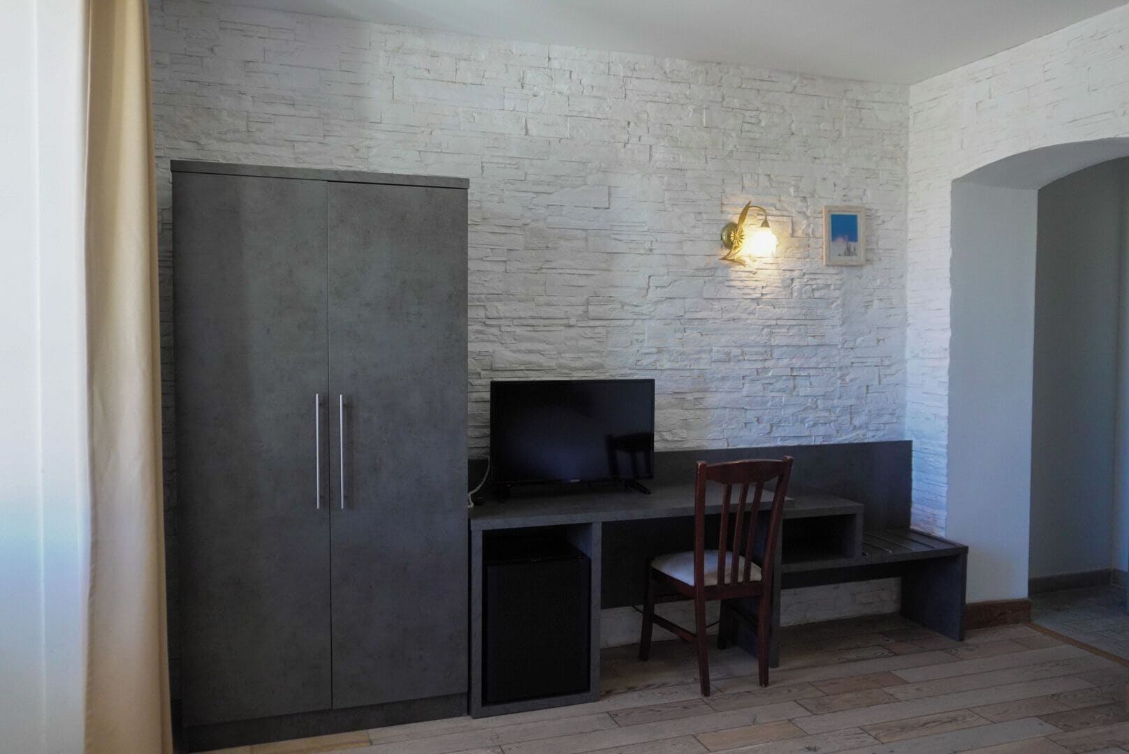 Une pièce avec un mur en pierre, un bureau avec une télévision, une chaise et une armoire.