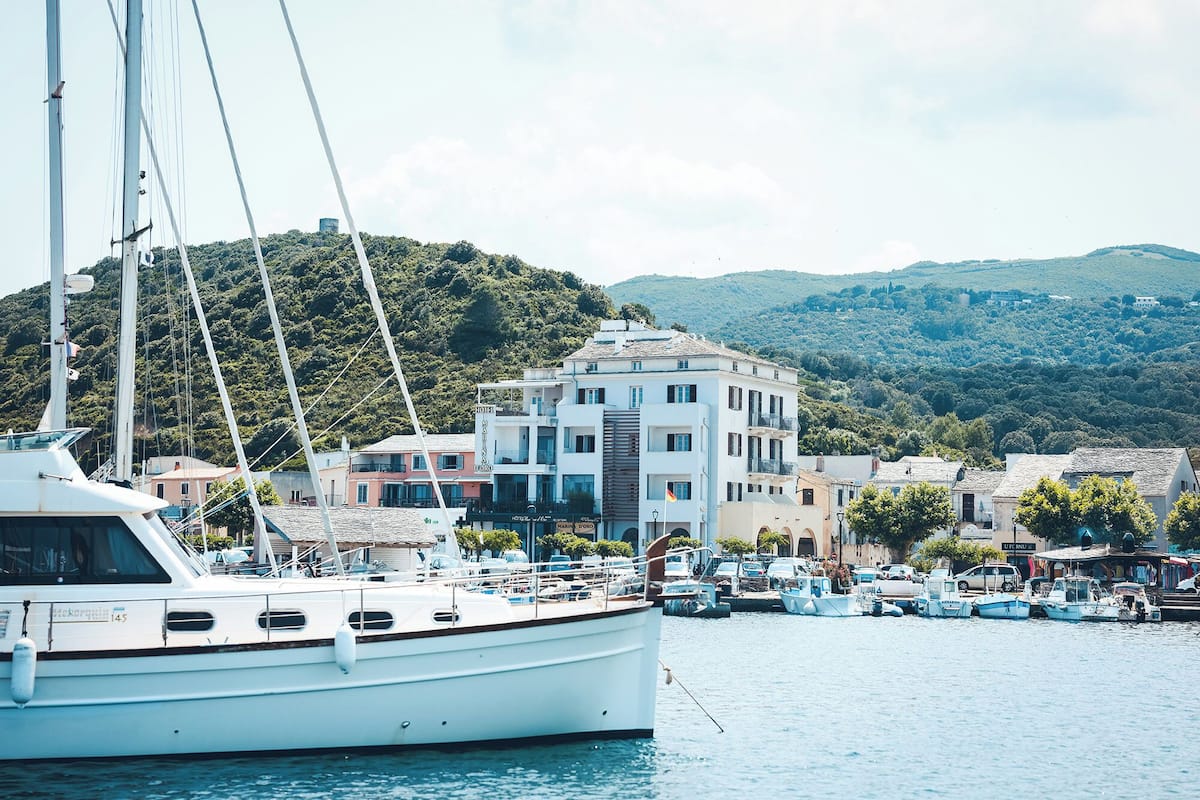 A marina with boats docked, a white building with multiple floors, and a hillside in the background.