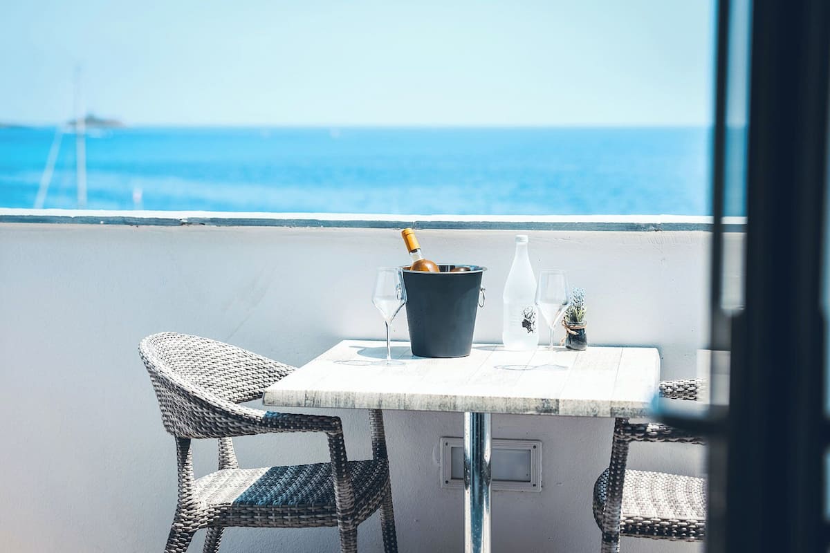 A balcony with a table, two wicker chairs, a bucket with a bottle and glasses, and a view of the sea.