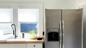 Moderni keittiö, jossa on valkoiset kaapit, ruostumattomasta teräksestä valmistettu jääkaappi, ikkuna, jossa on sälekaihtimet, ja puinen työtaso.