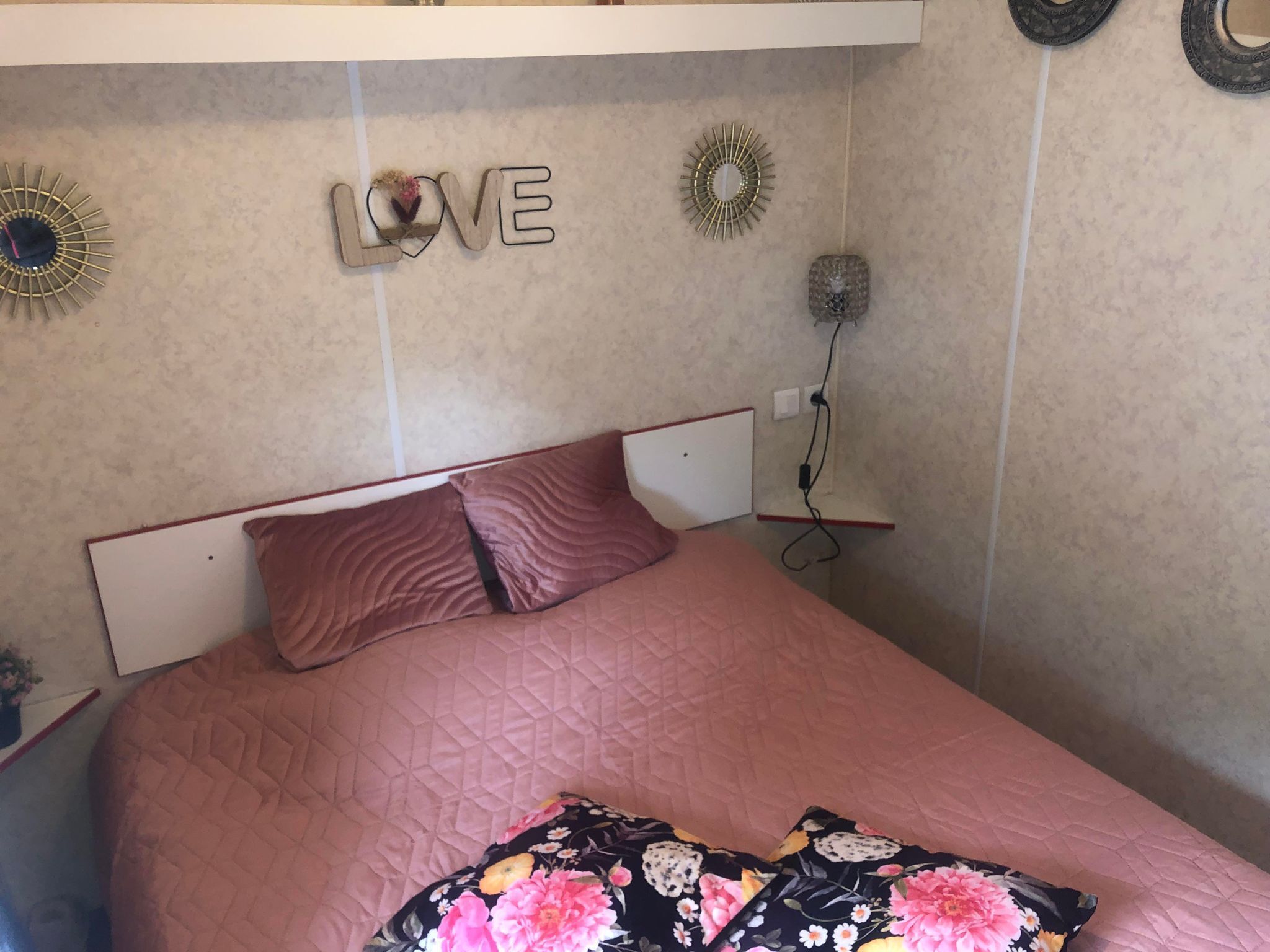 A neatly made bed with a pink quilted bedspread and floral pillows, a wall-mounted 'LOVE' decoration, and a small bedside table with a lamp.