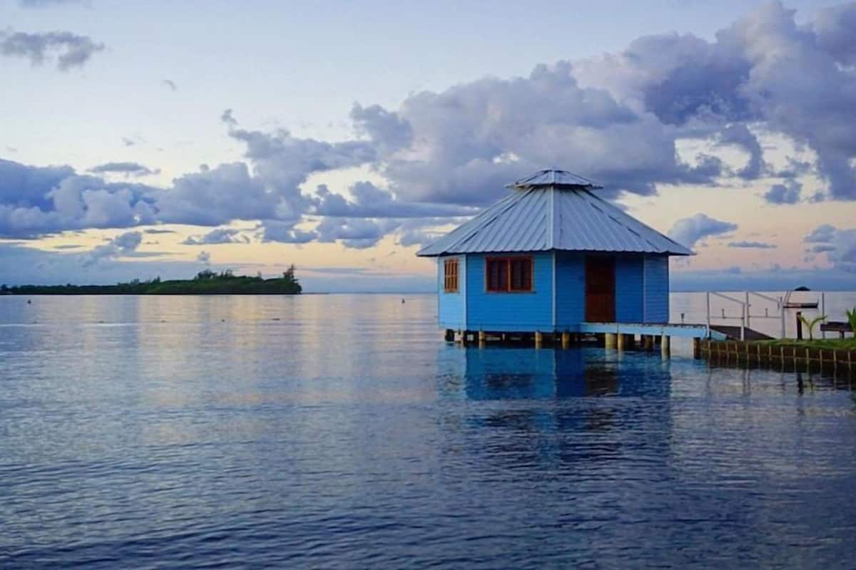 Uma casa azul com telhado de metal sobre pilares, acima de águas calmas, rodeada por um cais e um céu nublado.