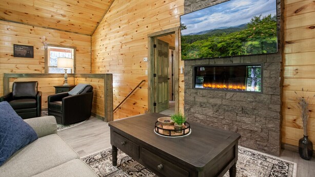 A cozy log cabin interior with a fireplace, wooden walls, a comfortable seating area, and a scenic view on the wall. - Allure Woods: View, HotTub, Outdoor Dining & Games (Sevierville)