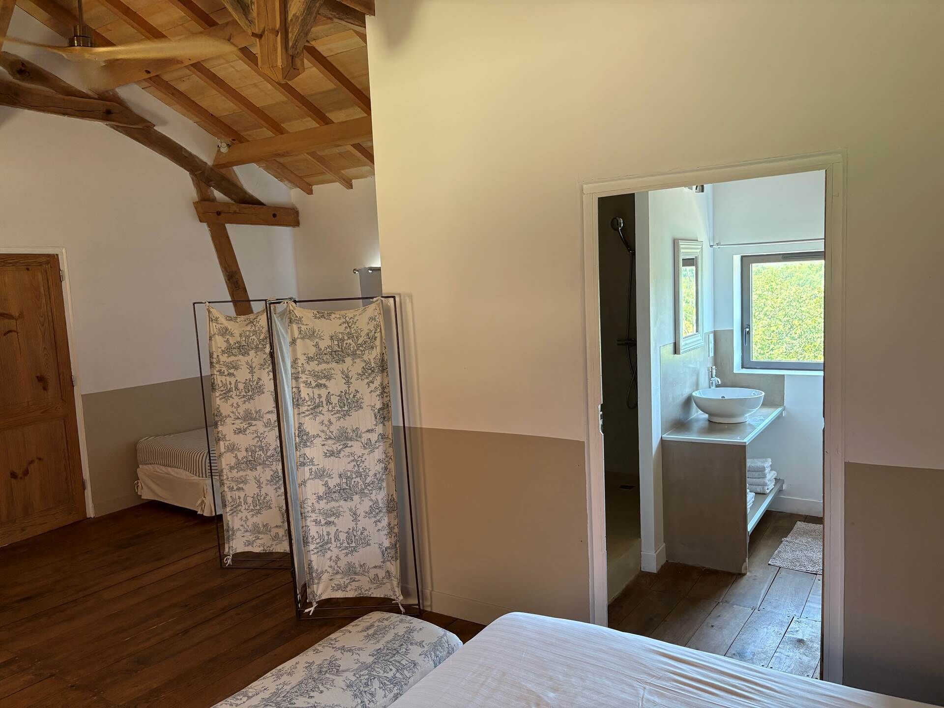 A bedroom with a wooden ceiling, a bed, a shower, and a bathroom with a sink and window.