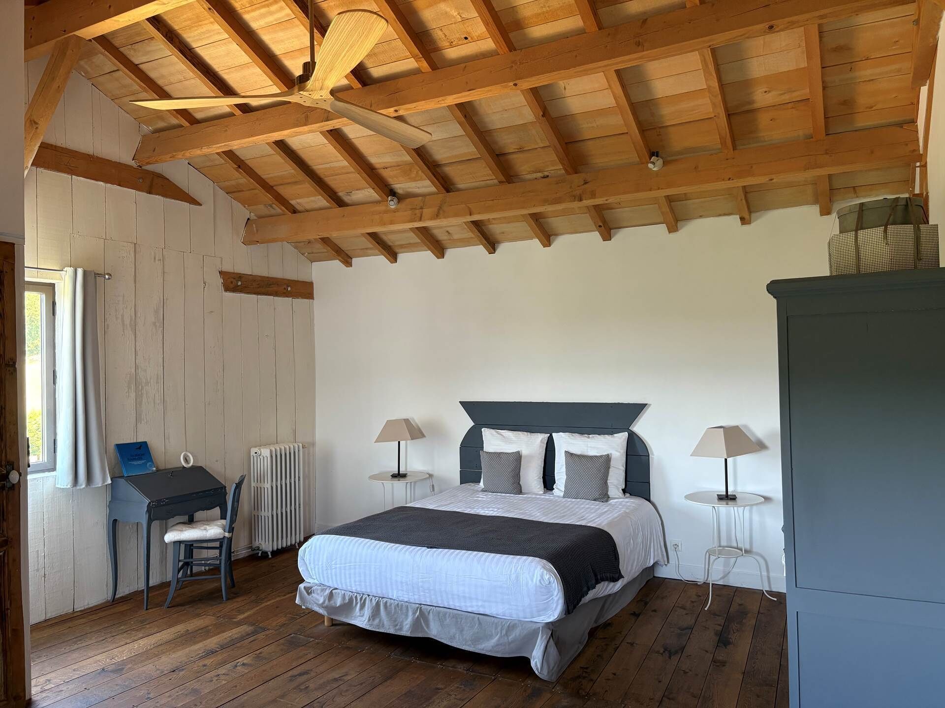 A bedroom with a wooden ceiling, a bed with a dark headboard, two bedside tables with lamps, a radiator, and a desk with a chair.