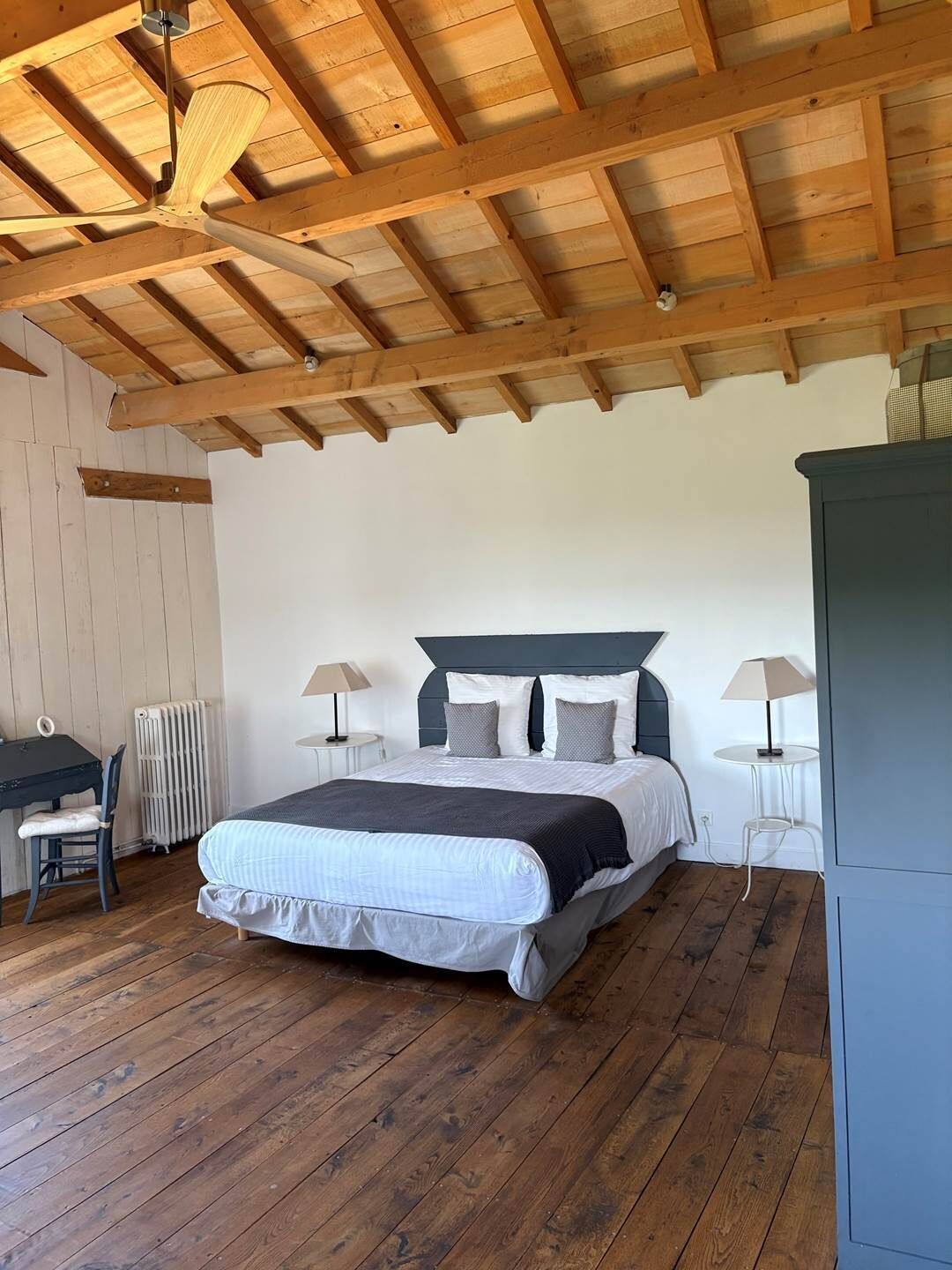 Quarto com teto de madeira, cama com cabeceira cinza, duas mesas de cabeceira com abajures, radiador e pequena escrivaninha com cadeira.
