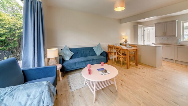 A modern living room with a blue sofa, a round table with two chairs, a dining area with a wooden table and chairs, and a kitchen with light-colored cabinets. - Discover coastal comfort with your perfect retreat in the heart of Beadnell. (Beadnell, near Chathill)
