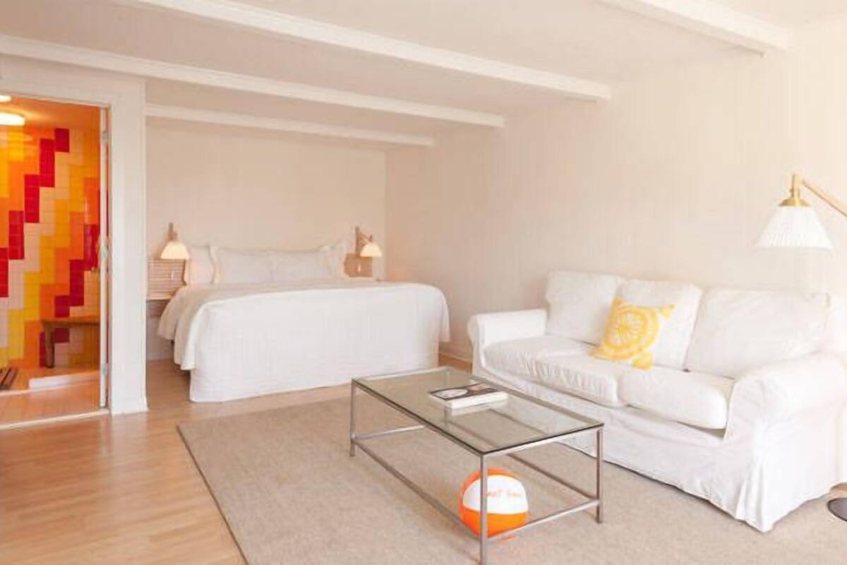 A modern hotel room with a large bed, a white sofa, a glass coffee table, and a lamp.