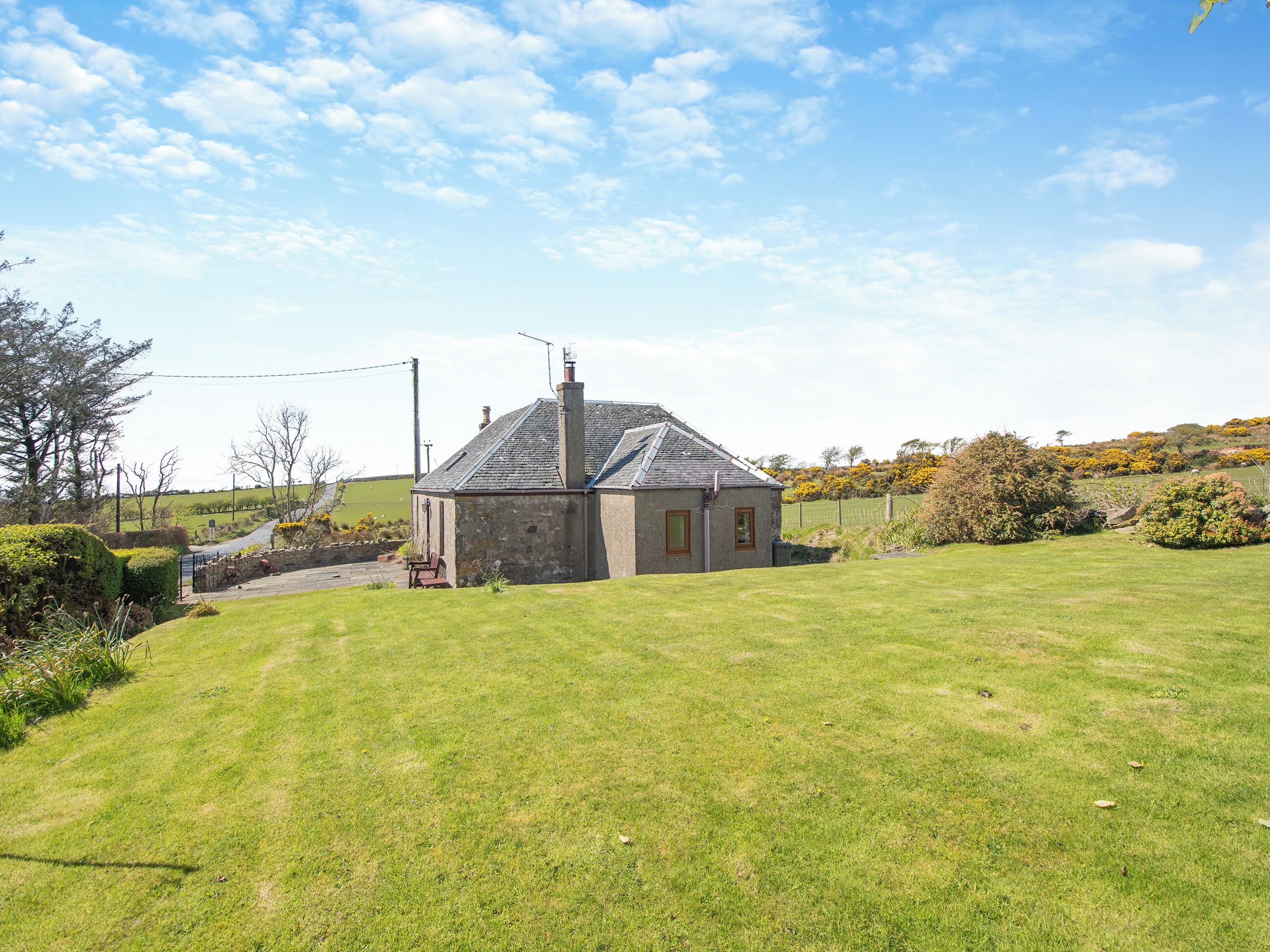A stone house with a sloped roof and a chimney, surrounded by a well-maintained lawn and a few trees.
