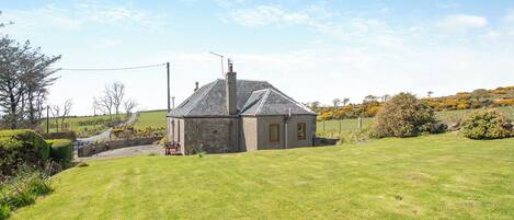 A stone house with a sloped roof and a chimney, surrounded by a well-maintained lawn and a few trees.