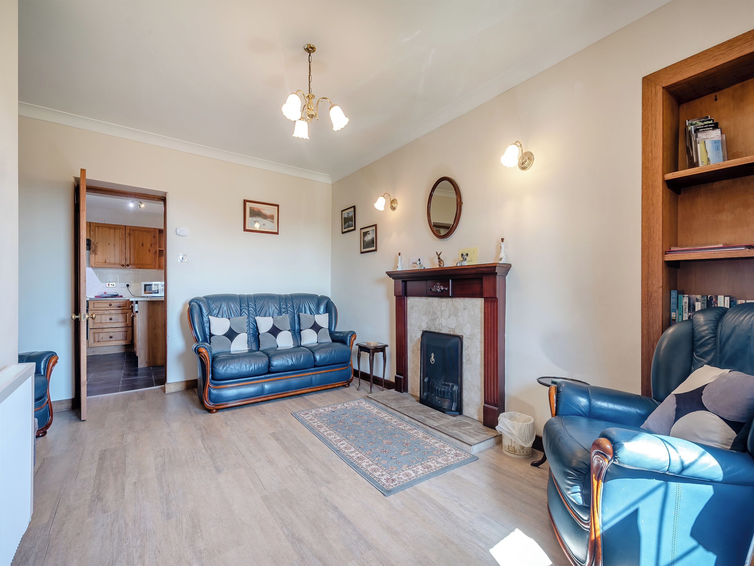A living room with a blue leather sofa, a fireplace, a wooden bookshelf, and a kitchen visible through an open door.