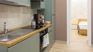 Une cuisine compacte équipée d’un évier, d’une machine à café et d’un grille-pain. Elle comprend un plan de travail en bois et un four fixé au mur. La cuisine donne sur une chambre à coucher avec un lit et une table de chevet.
