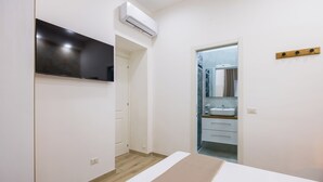 Une chambre à coucher comprenant un lit, un téléviseur, un climatiseur, une salle de bain avec un lavabo et un miroir, et une étagère en bois.
