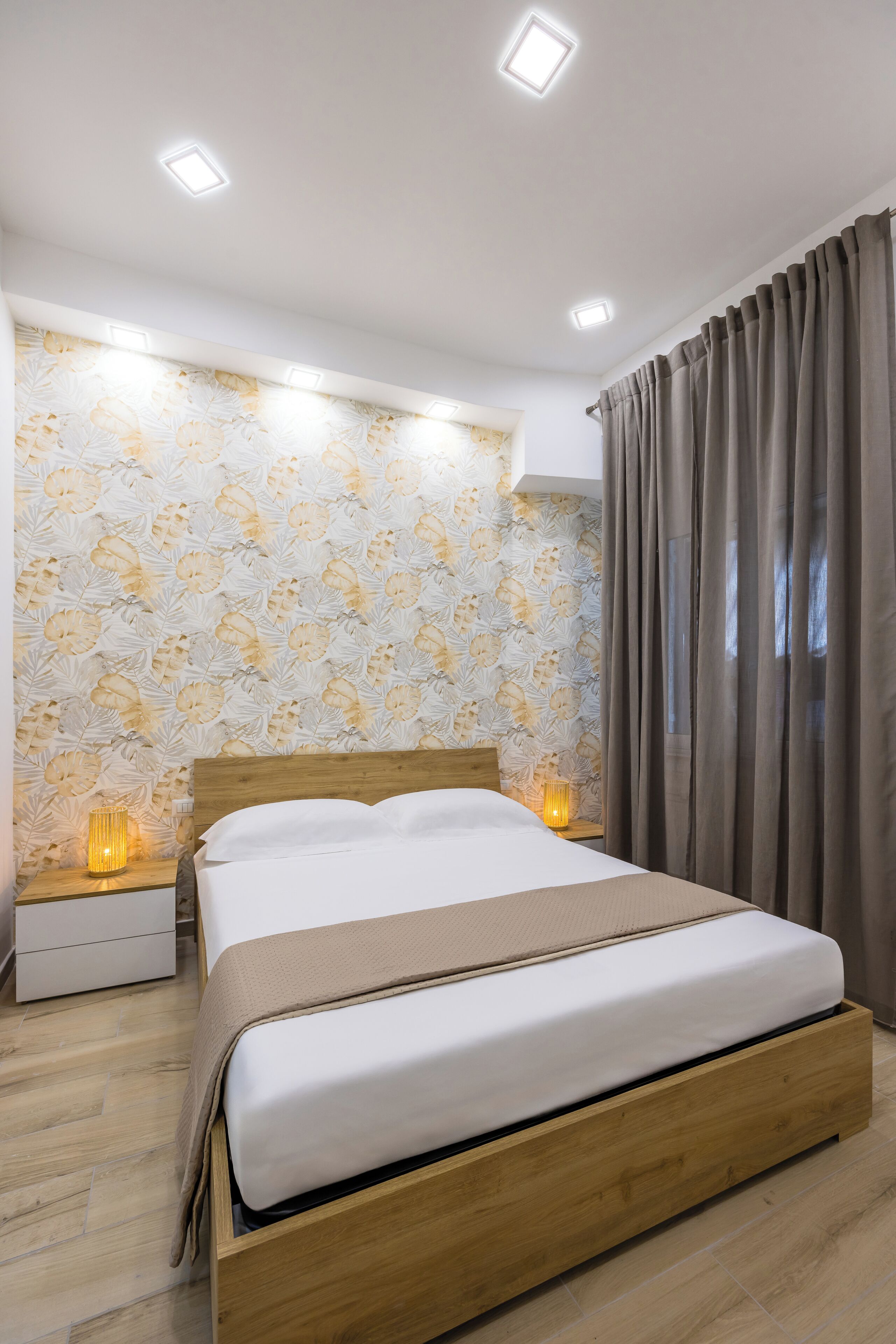 Una camera da letto moderna con un letto in legno, biancheria da letto bianca, carta da parati floreale e un comodino con una lampada.