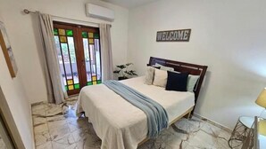 Een slaapkamer met een bed, een 'WELCOME' bord, een potplant en uitzicht door een deur met gordijnen.