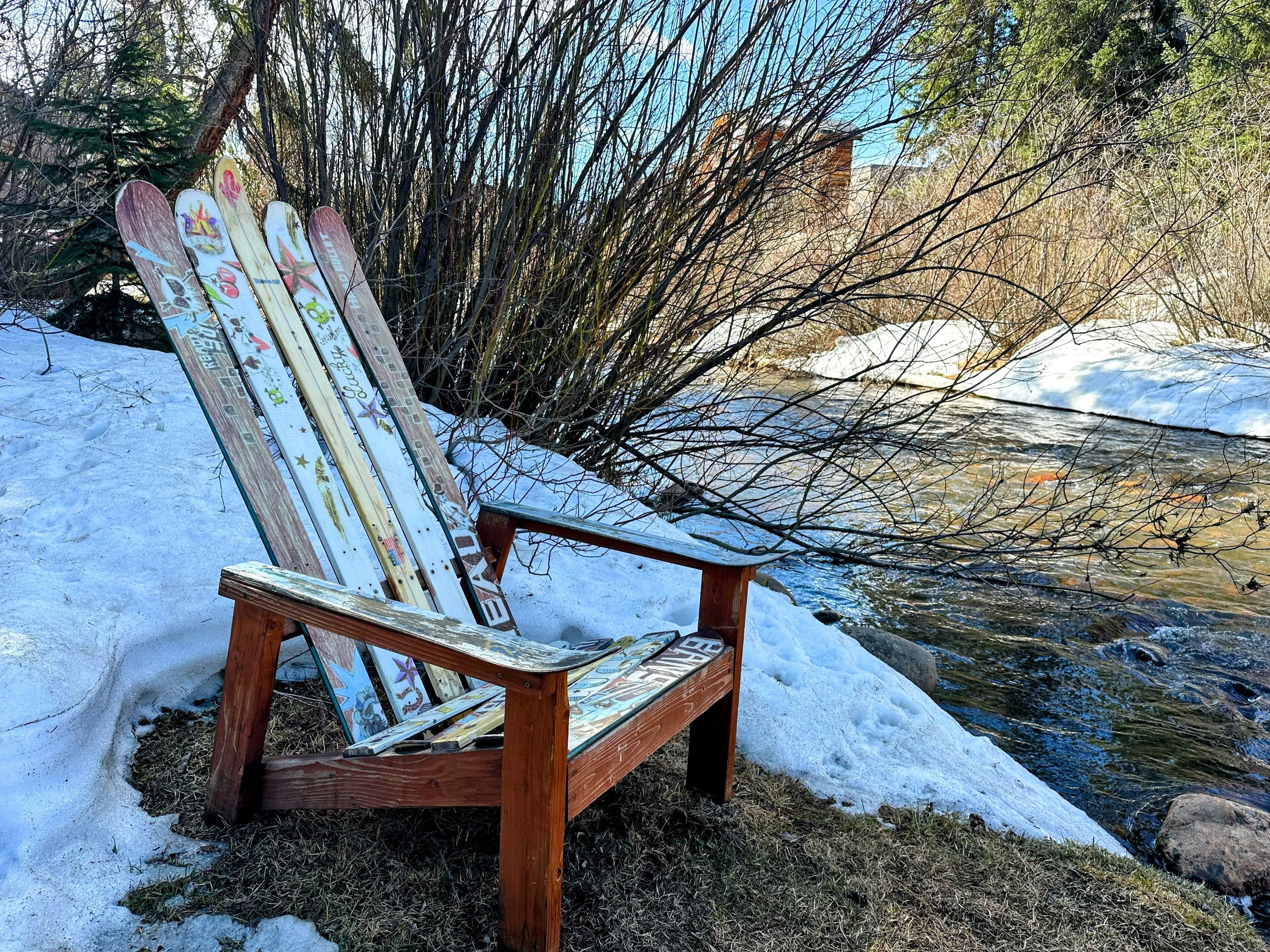 Una poltrona Adirondack di legno con decorazioni colorate, posizionata vicino a un sentiero innevato e un ruscello.