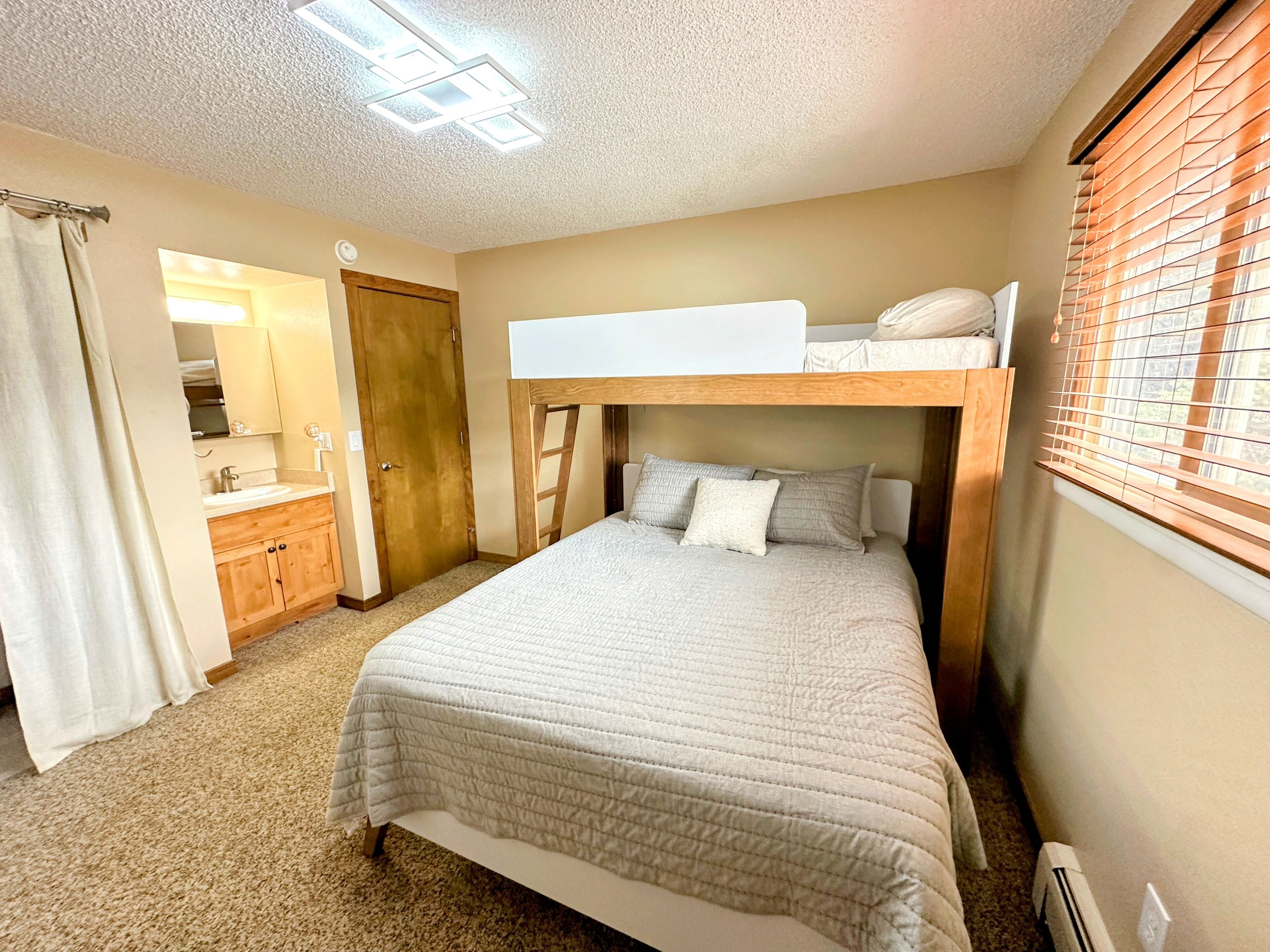 A compact bedroom with a bunk bed, a single bed, a wooden wardrobe, a bathroom with a mirror, and a window with blinds.