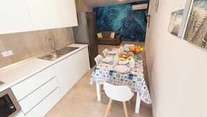 A compact kitchen with white cabinets, a stainless steel sink, and a built-in oven. A dining area with a table set for two, featuring a patterned tablecloth and a fruit basket. - Altamarea Sea Apartments (Balestrate)