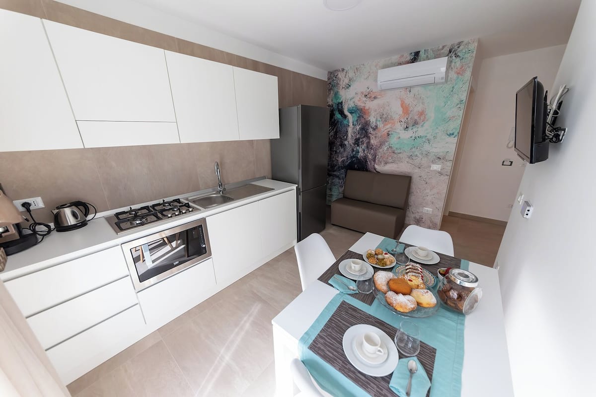A compact kitchen with a gas stove, sink, and built-in oven. A dining area with a table set for two, featuring a variety of pastries and a glass of water.
