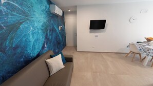 A living room with a blue abstract wall, a flat-screen TV mounted on the wall, a dining table with chairs, and a sofa with a cushion. - Altamarea Sea Apartments (Balestrate)