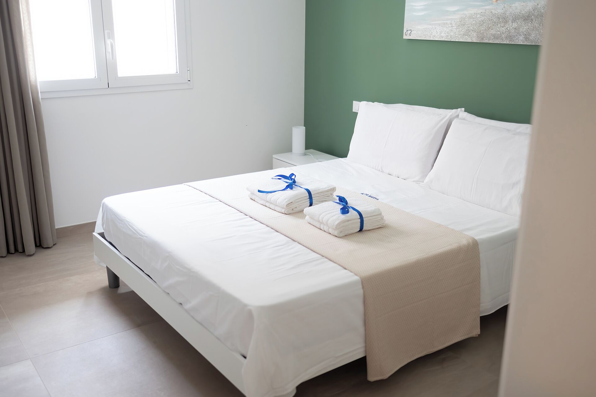 Una cama bien hecha con sábanas blancas y dos toallas dobladas atadas con cintas azules, una mesita de noche blanca con una lámpara, y una pared verde con un cuadro.