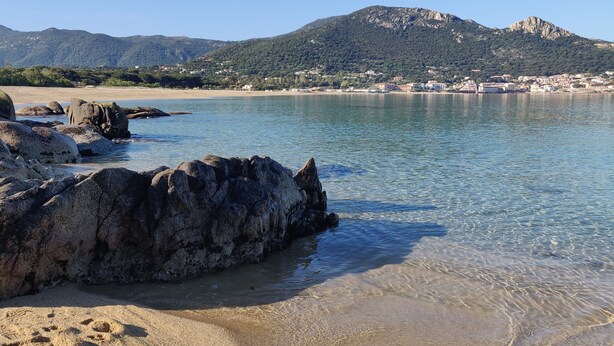 Una spiaggia con grosse rocce e acqua cristallina, una cittadina in lontananza e montagne sullo sfondo.