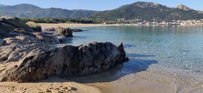 Une plage avec de gros rochers et une eau cristalline, une ville lointaine et des montagnes en arrière-plan.