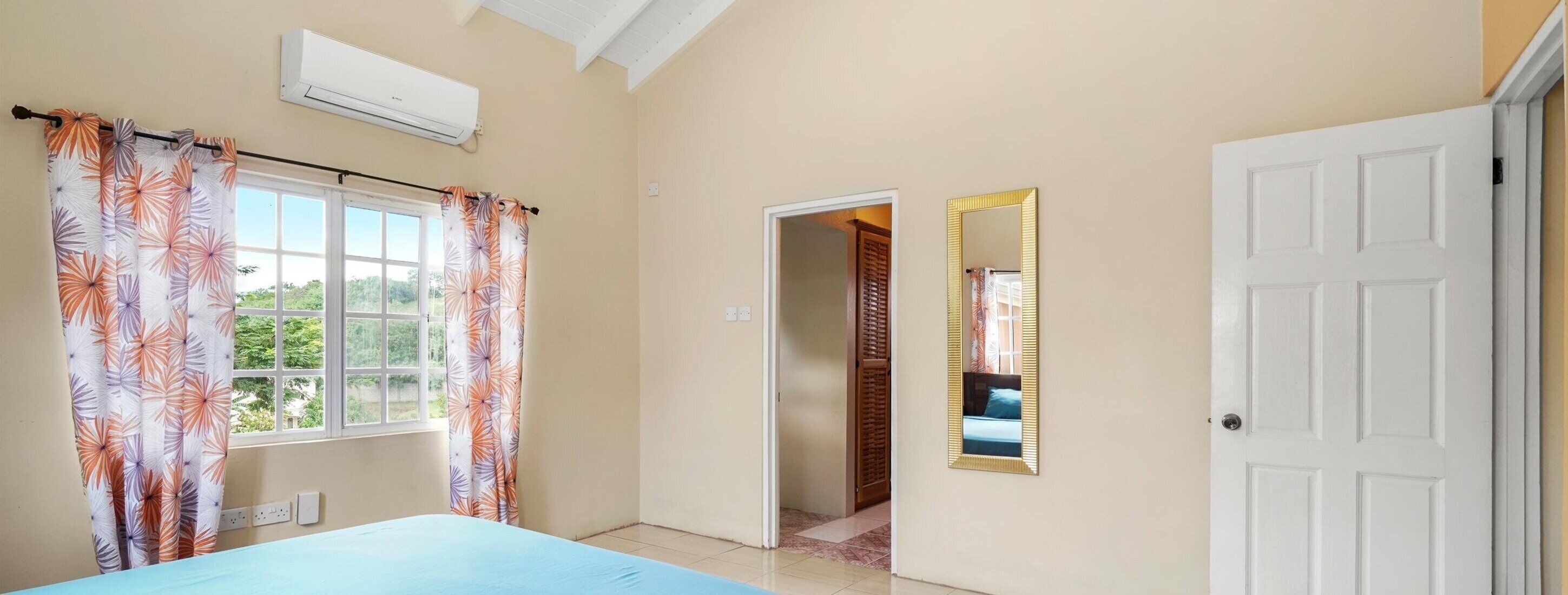 Une chambre à coucher avec un lit, une fenêtre avec des rideaux, un appareil de climatisation, une porte et un miroir.