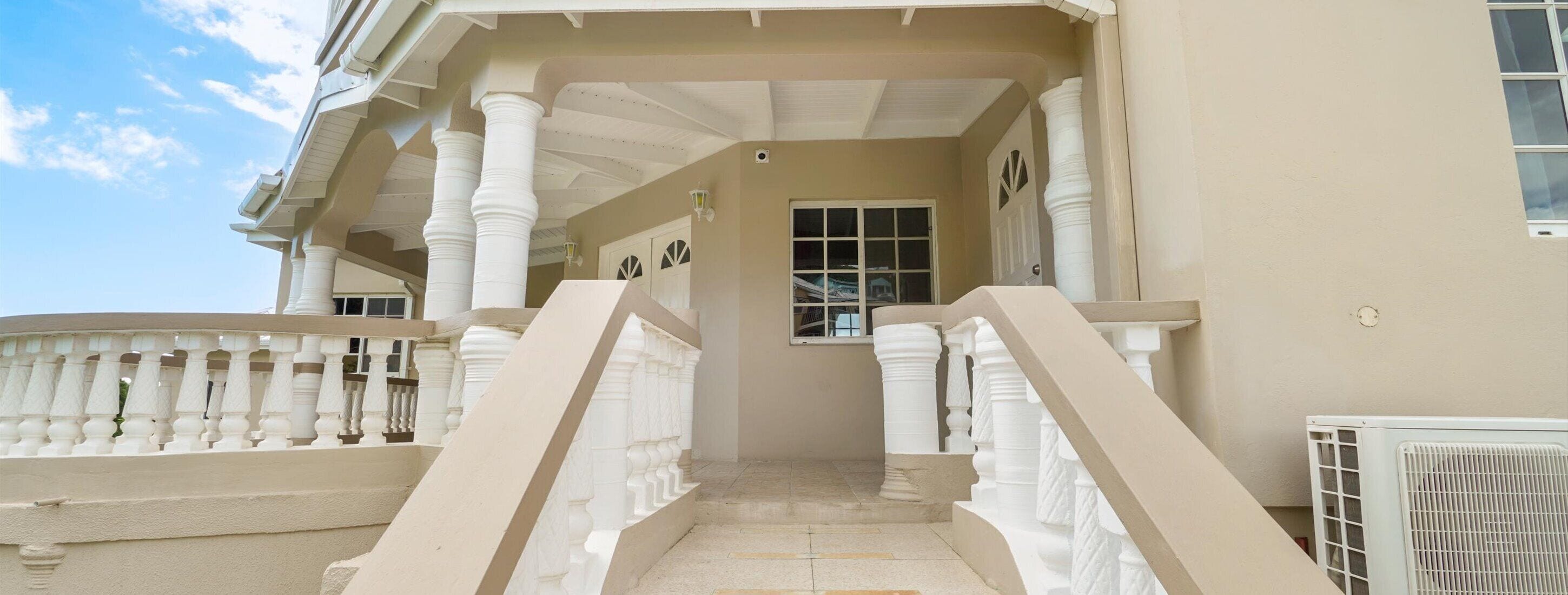 Un escalier menant à l’entrée d’un bâtiment, avec des balustrades et des colonnes blanches, un ciel dégagé et une fenêtre offrant une vue.