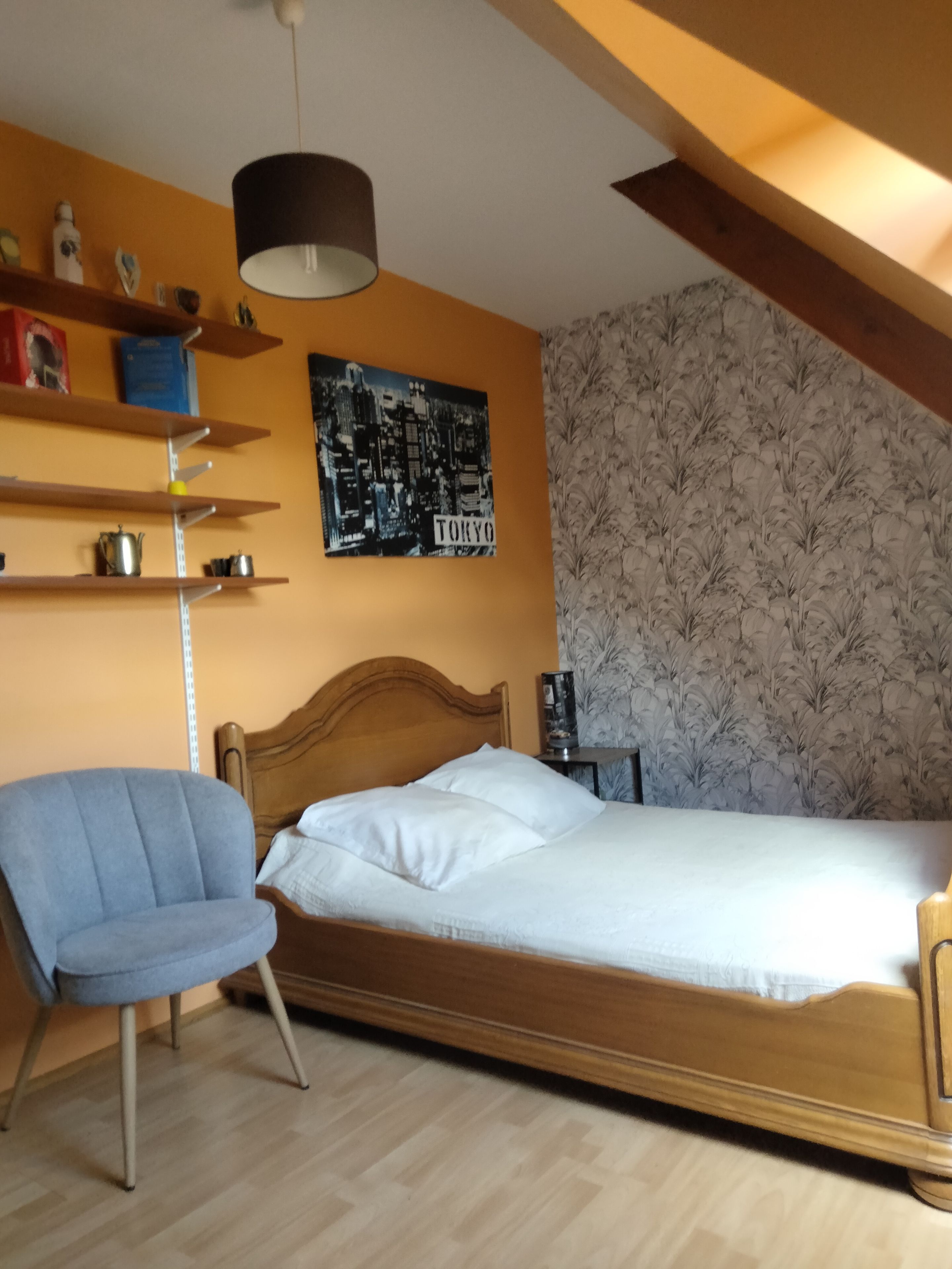 Un dormitorio con una cama de madera, una silla azul, una estantería con objetos, un cartel con un motivo urbano y un papel tapiz estampado.