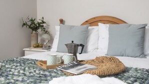 Ein ordentlich bezogenes Bett mit einem Kissen mit Punkten, ein hölzernes Kopfende, ein Nachttisch mit Pflanze, eine Kaffeemaschine und ein Tablett mit Zeitschrift und Tassen.