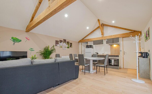 A modern living room with a grey sofa, a dining area with a round table and chairs, and a kitchen with wooden beams and white cabinets.