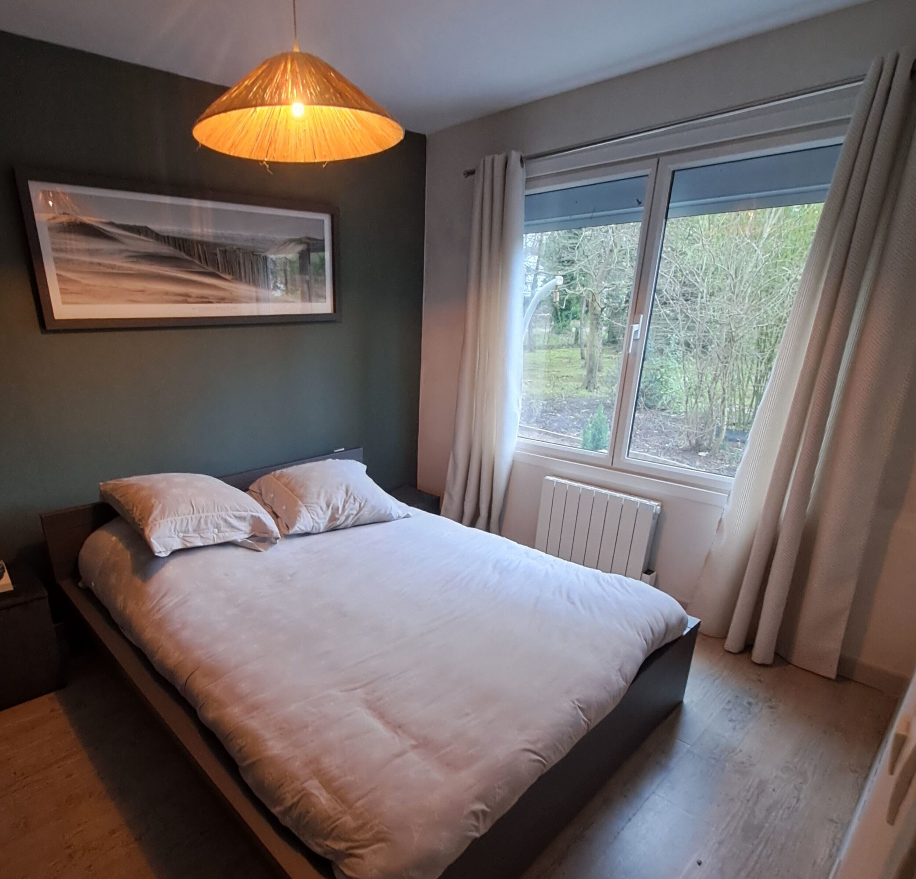 A bedroom with a bed, a hanging light fixture, a window with curtains, and a radiator.