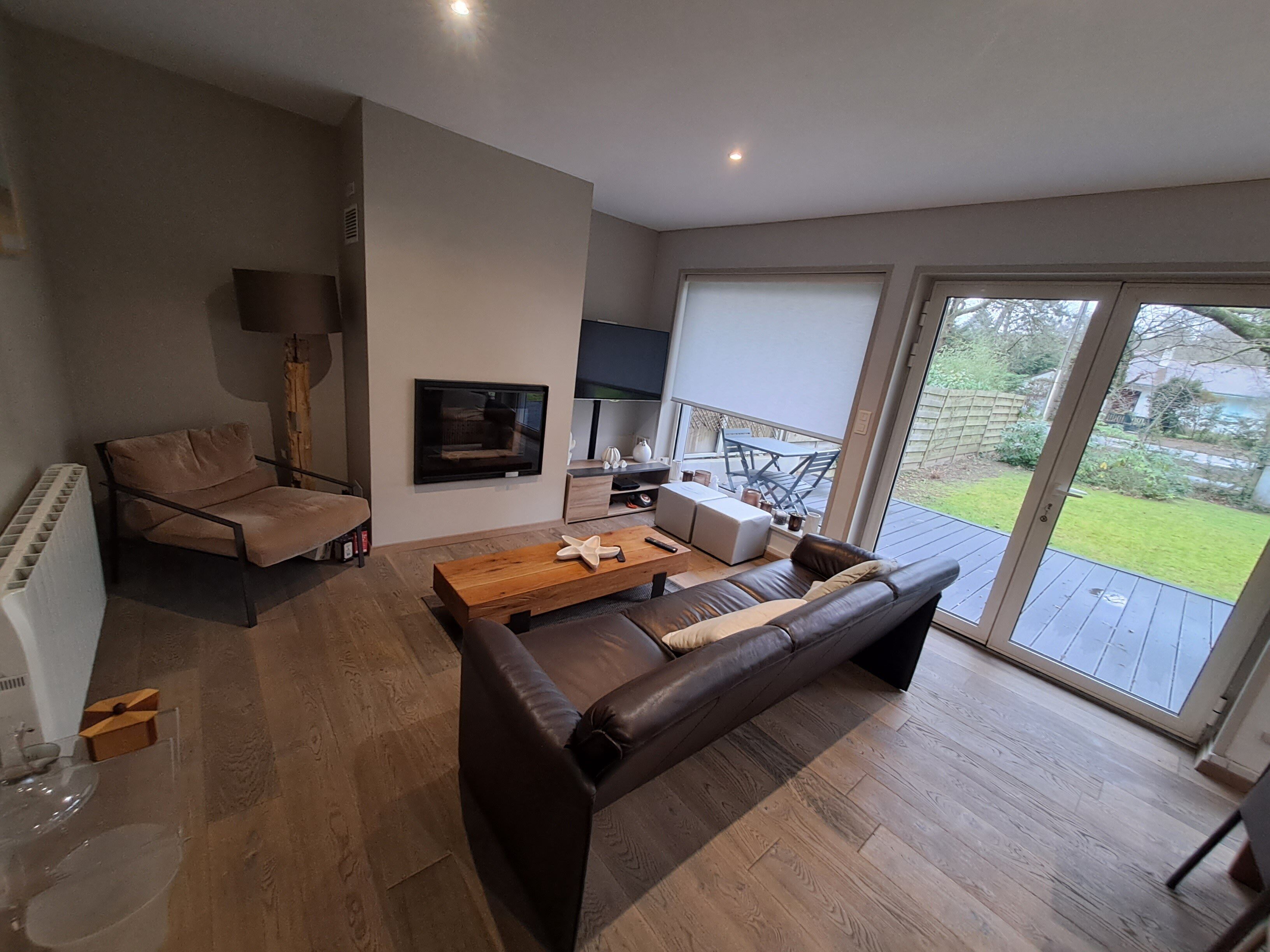 A modern living room with a sofa, a wooden coffee table, and a TV mounted on the wall. There is a sliding glass door leading to a patio with outdoor furniture.