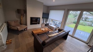 A modern living room with a sofa, a wooden coffee table, and a TV mounted on the wall. There is a sliding glass door leading to a patio with outdoor furniture. - Villa touquettoise near the sea (Le Touquet-Paris-Plage)