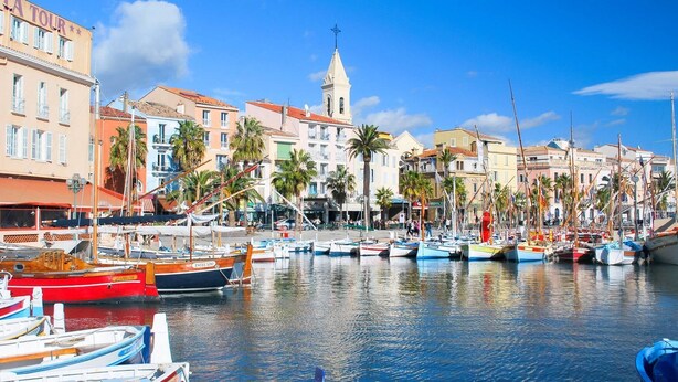 Un puerto deportivo con varias embarcaciones amarradas, una iglesia con un campanario y edificios coloridos a lo largo del paseo marítimo.
