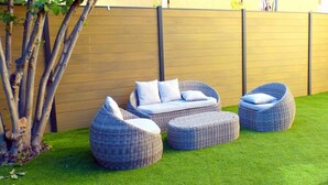 Un espacio exterior con mobiliario de mimbre, incluyendo un sofá y dos sillones, frente a una valla de madera y un árbol.