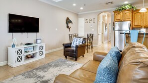 Een moderne woonkamer met een leren bank, een aan de muur bevestigde televisie, een eethoek met stoelen en een keuken met houten kastjes.