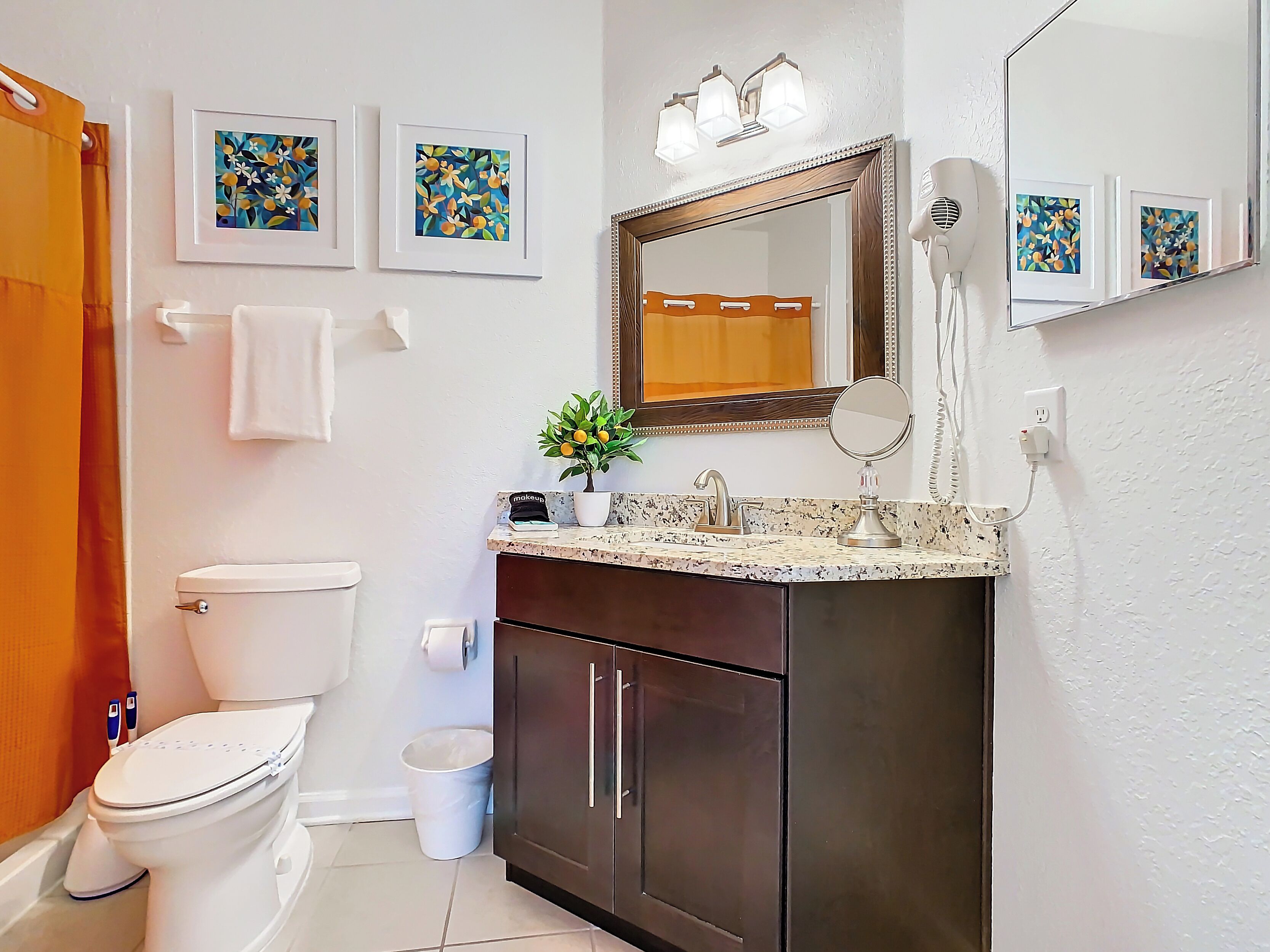 A bathroom with a toilet, sink, vanity, and a mirror. There are framed floral artworks on the wall, a potted plant on the sink, and a towel hanging on the wall.