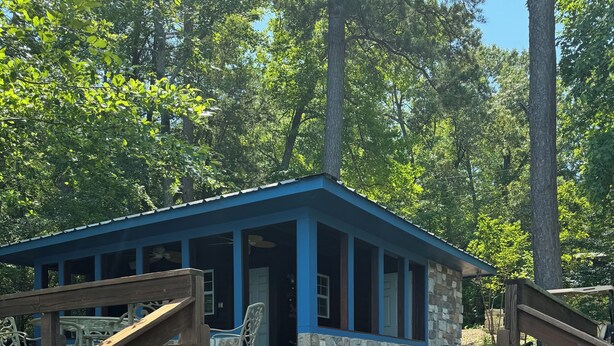 A wooden deck with a table and chairs, leading to a stone and blue-roofed building with a covered porch.