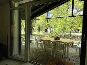 A covered outdoor area with a table and chairs, a basket, and a view of a wooded area. - Maison Bruyères by Agence Bru - 483473 (Lacanau)