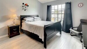 A bedroom with a bed, a nightstand, a chair, a ceiling fan, and a window with curtains. - Disney Themed Condo mins from Disney attractions (Kissimmee)