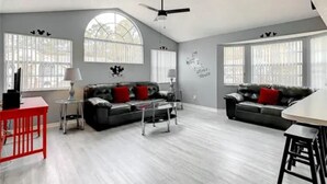 A spacious living room with two sofas, a coffee table, and a red table. There are large windows with blinds and a ceiling fan. - Disney Themed Condo mins from Disney attractions (Kissimmee)