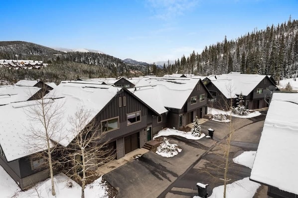 A cluster of modern cabins with sloped roofs, surrounded by snow-covered ground and trees, set against a backdrop of mountains and clear sky.